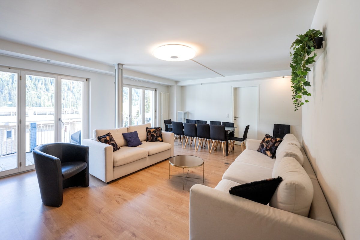 A spacious living area is displayed, featuring a large light-colored sofa with accent pillows and a round glass coffee table. A dark chair is positioned nearby. The room is filled with natural light from floor-to-ceiling windows, creating a comfortable and open atmosphere.