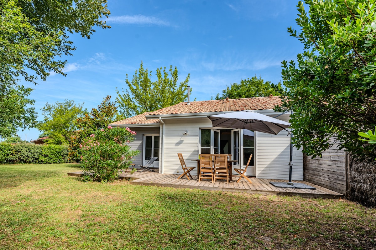 An outdoor wooden terrace is pictured, furnished with a large table and several chairs. A large parasol provides shade. The surrounding area features well-maintained grass and a variety of greenery, offering a sense of privacy and tranquility.