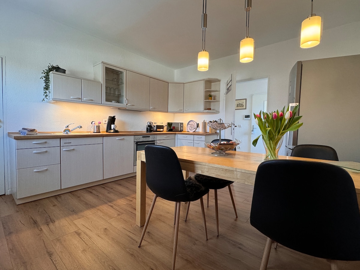The kitchen area features light wood cabinetry and modern appliances. A central wooden table is surrounded by four black chairs. A vase with tulips is placed on the table, and warm lighting enhances the inviting space.
