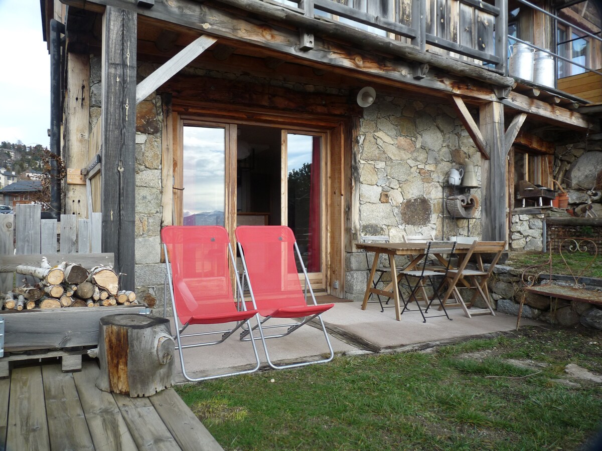 An outdoor seating area features two red lounge chairs positioned on a patio. A small wooden table is set nearby, surrounded by natural stone walls and rustic wooden beams. A stack of firewood is neatly arranged to the side, enhancing the cozy outdoor space.