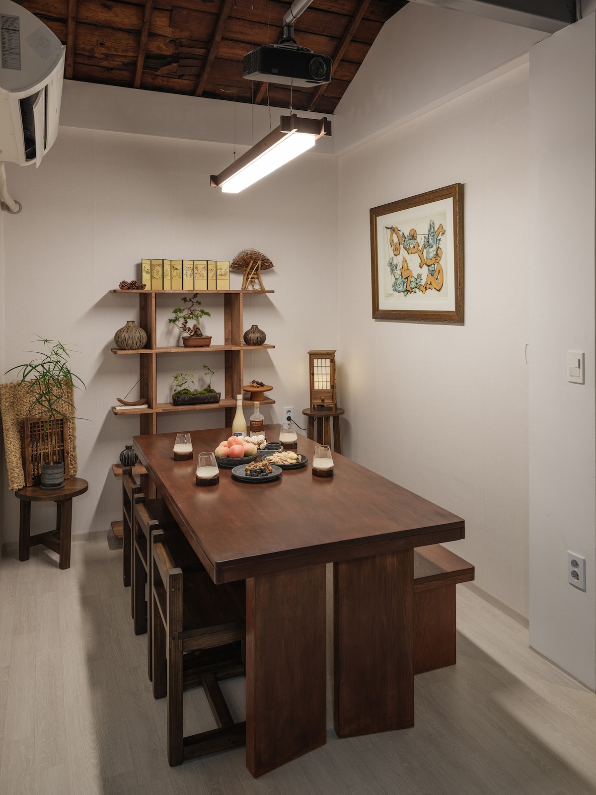 A dining area is presented, featuring a wooden table surrounded by several smaller chairs. A traditional Korean painting is displayed on the wall, while decorative items are arranged on a shelf, including plants and ceramics. Soft lighting is provided by overhead fixtures.