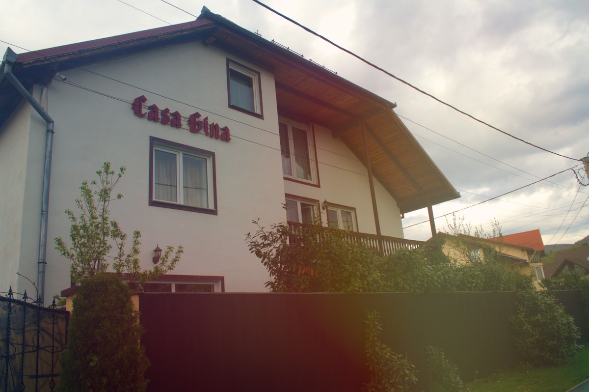 The exterior of Casa Gina is shown, featuring a two-story white building with a brown roof. The property is set in a tranquil area, surrounded by greenery and a black fence. The name 'Casa Gina' is displayed prominently on the front facade.