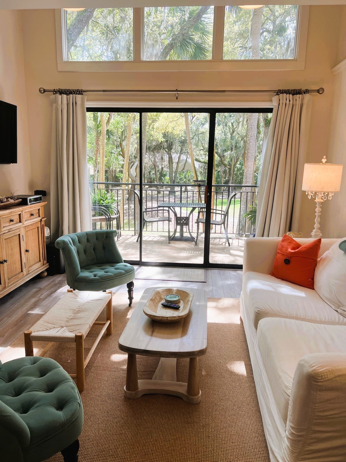 A cozy living area features light, airy curtains framing sliding glass doors that lead to a private outdoor space. A white sofa is complemented by two accent chairs in a soft green hue. A natural wood coffee table is positioned centrally on a woven rug.