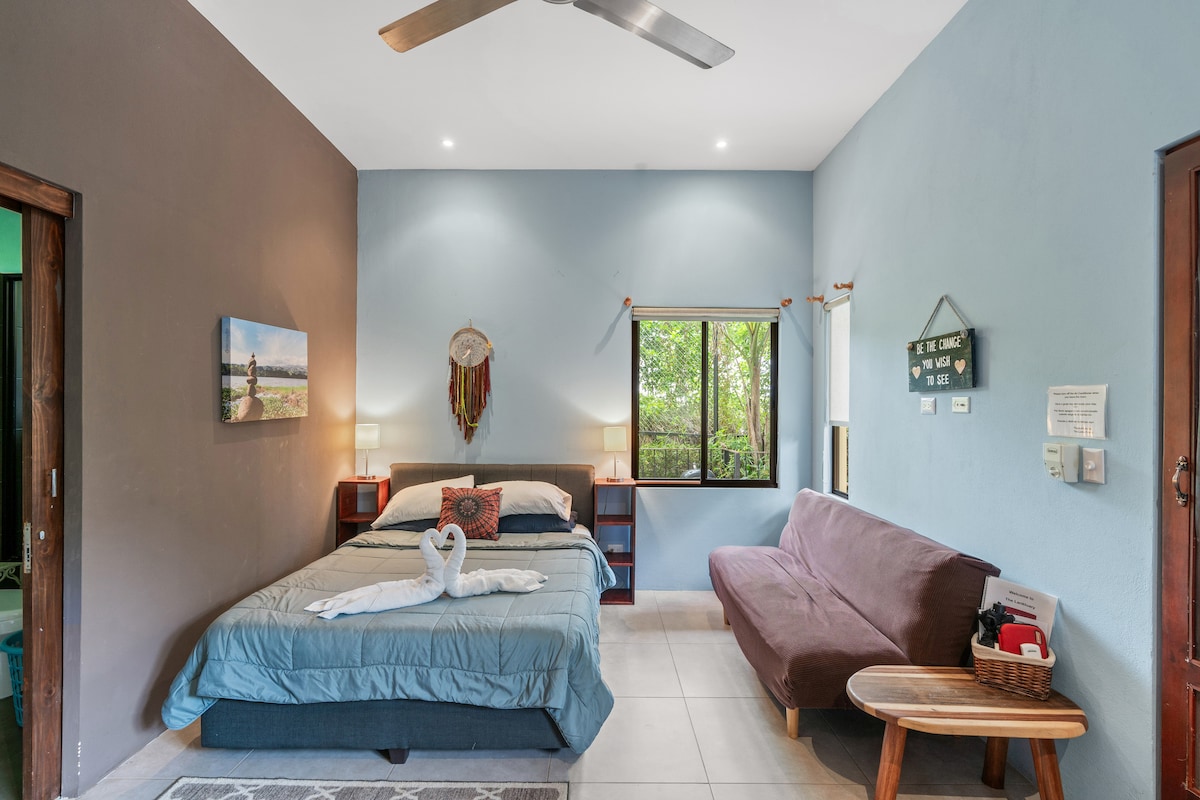 A cozy studio room is displayed, featuring a queen-sized bed with a neatly arranged blanket and soft pillows. A sofa bed and a wooden table are visible. Natural light enters through a window framed by greenery, complementing the peaceful ambiance of the space.