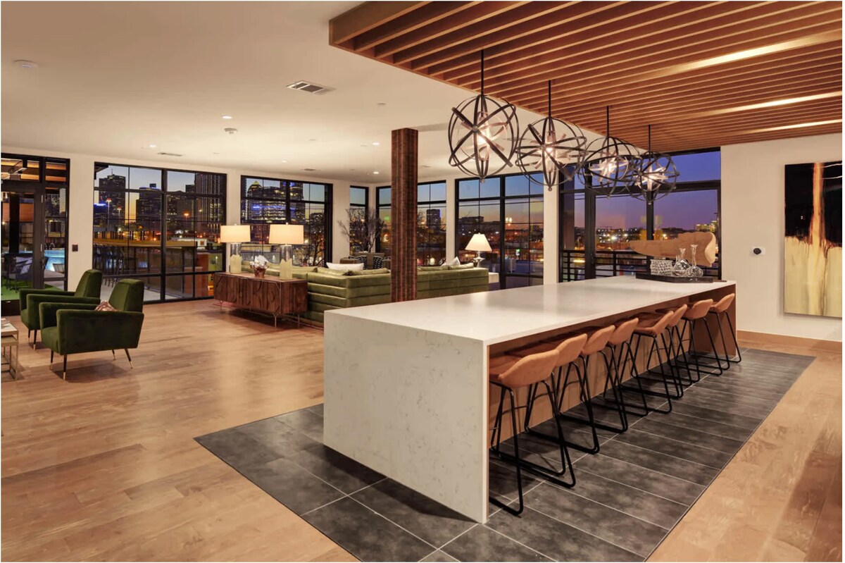 A modern communal space features a long white countertop bar with high stools, illuminated by stylish pendant lights. Large windows offer views of the city skyline, and comfortable green armchairs are arranged nearby. The wooden ceiling and warm lighting create an inviting environment.
