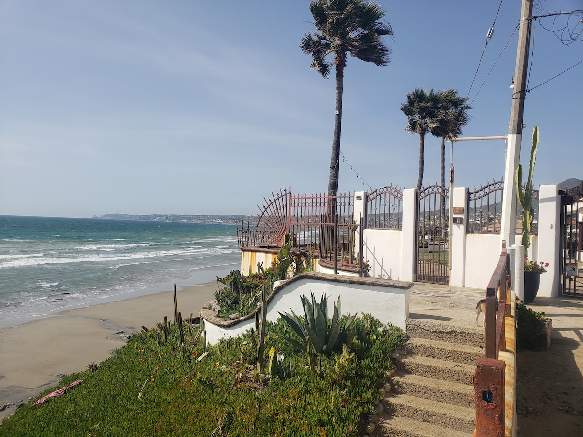 A gated entrance is visible, leading toward the beach. Lush greenery and cacti are planted alongside the path, which descends toward the sandy shore. Palm trees sway gently in the breeze, while the ocean waves can be seen in the background under a clear sky.