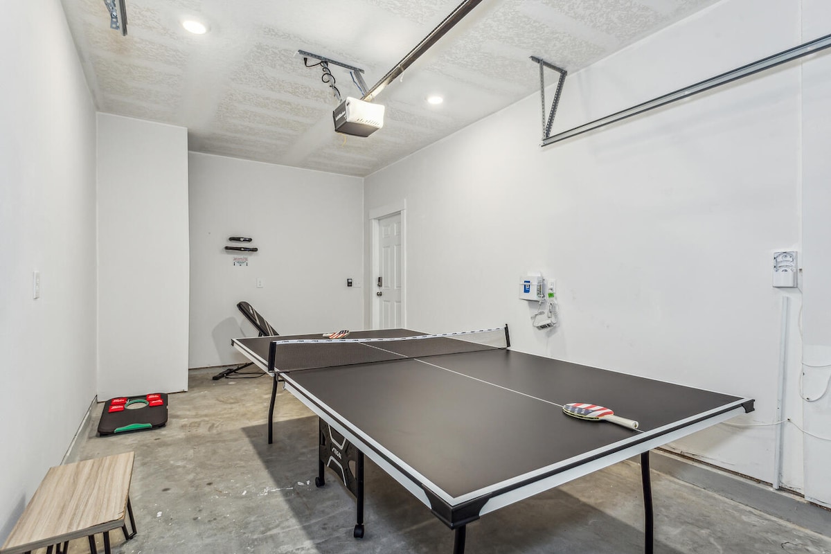 A gameroom features a black ping pong table positioned centrally on a concrete floor, with a small wooden bench nearby. A wall mount projector is visible above the table, and there is ample natural light coming from an unobstructed window.