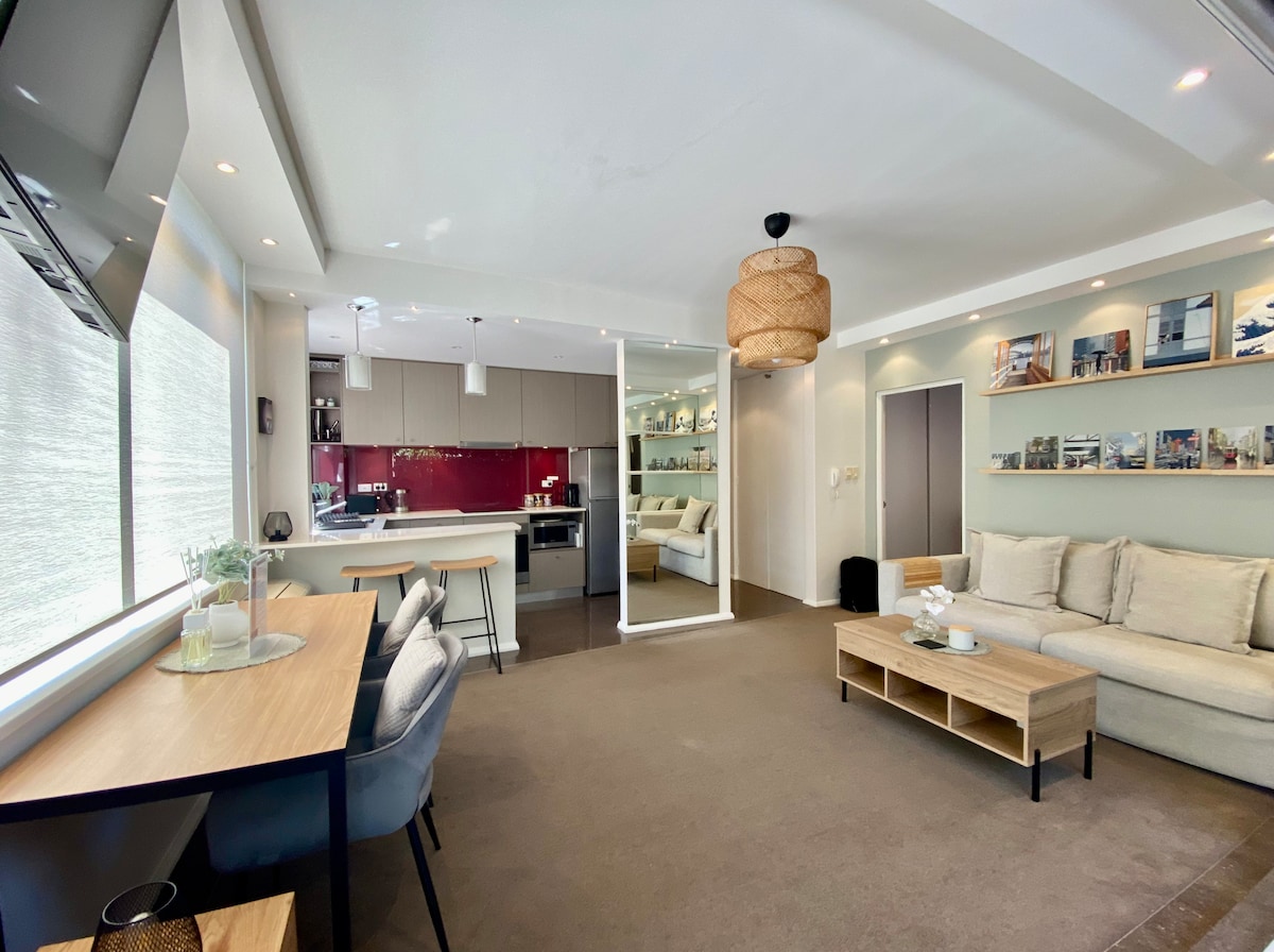 A bright and modern living space features a large, comfortable sectional sofa with light cushions. A sleek wooden coffee table sits in front, with a stylish workspace area showcasing a desk and chairs. The contemporary kitchen is visible in the background, accented by red cabinetry.