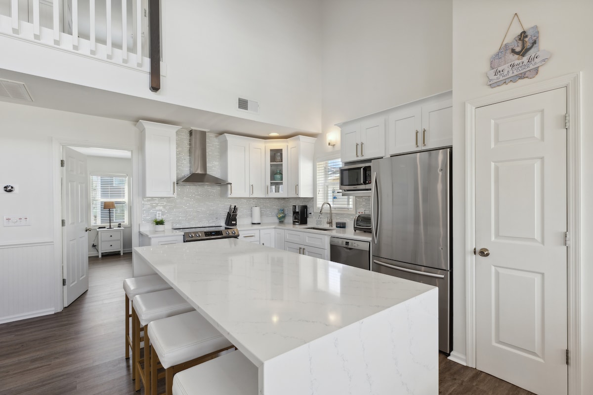 A spacious kitchen is shown, featuring a large central island with seating for four. Stainless steel appliances and a sleek backsplash complement the quartz countertops. Natural light fills the area through nearby windows, enhancing the inviting and functional layout.