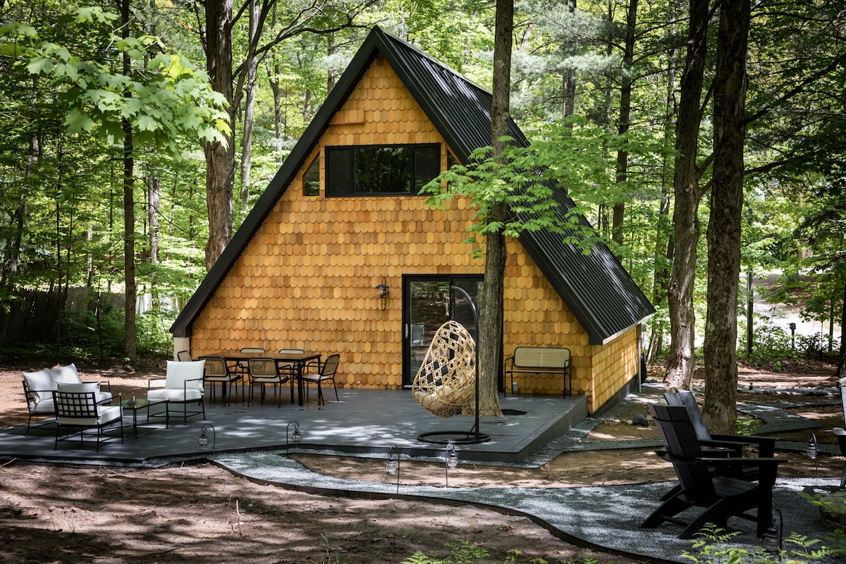 The A-Frame cabin showcases a landscaped exterior surrounded by lush trees. A spacious deck features a dining table with chairs and a cozy hanging seat. Comfortable seating areas are complemented by shaded sections, accentuating the serene woodland setting.