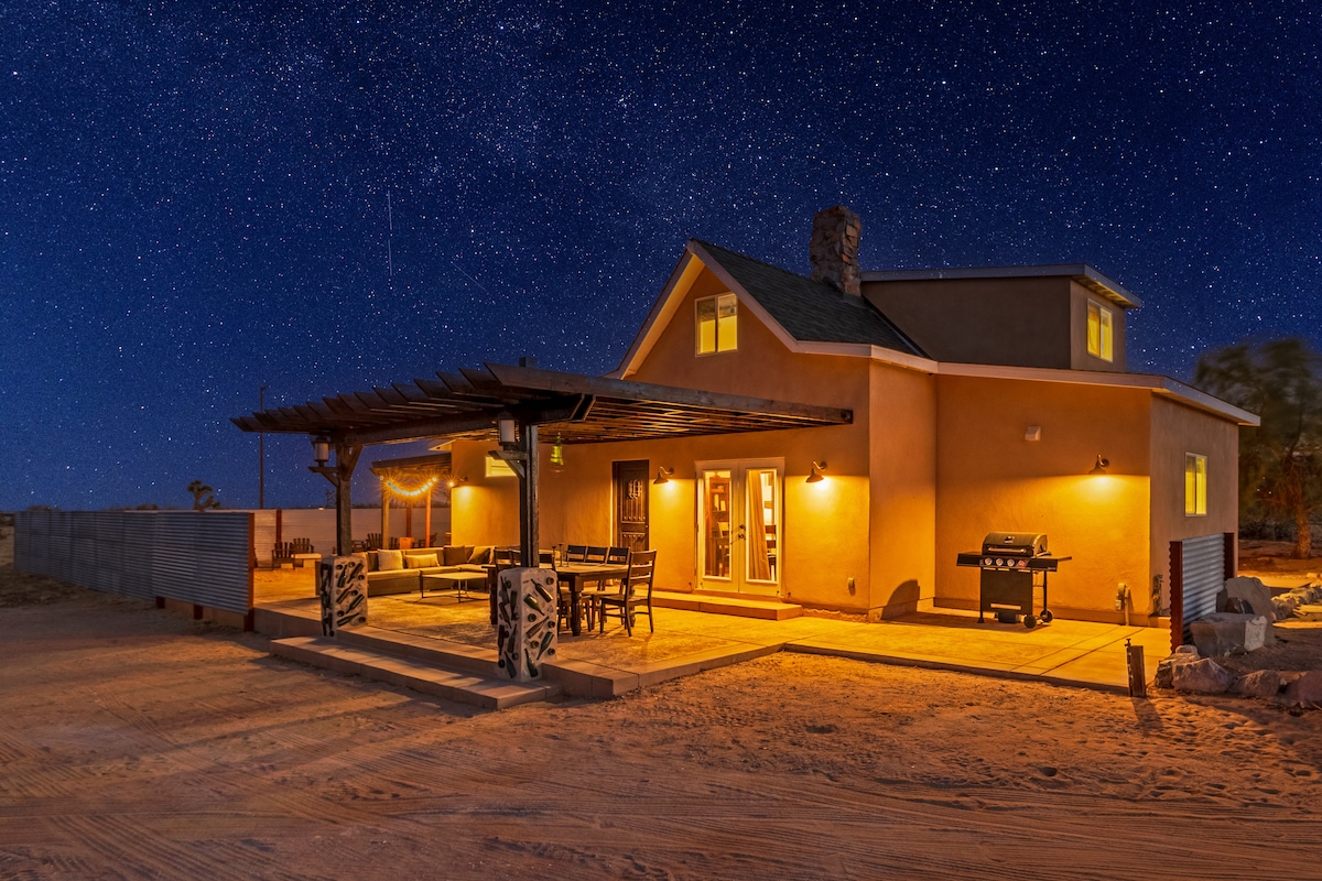 A modern desert retreat is illuminated under a starry night sky, featuring a spacious patio area with outdoor seating and a barbecue. The structure showcases large windows, enhancing the inviting ambiance. The surrounding landscape reveals a sandy surface typical of desert terrain.