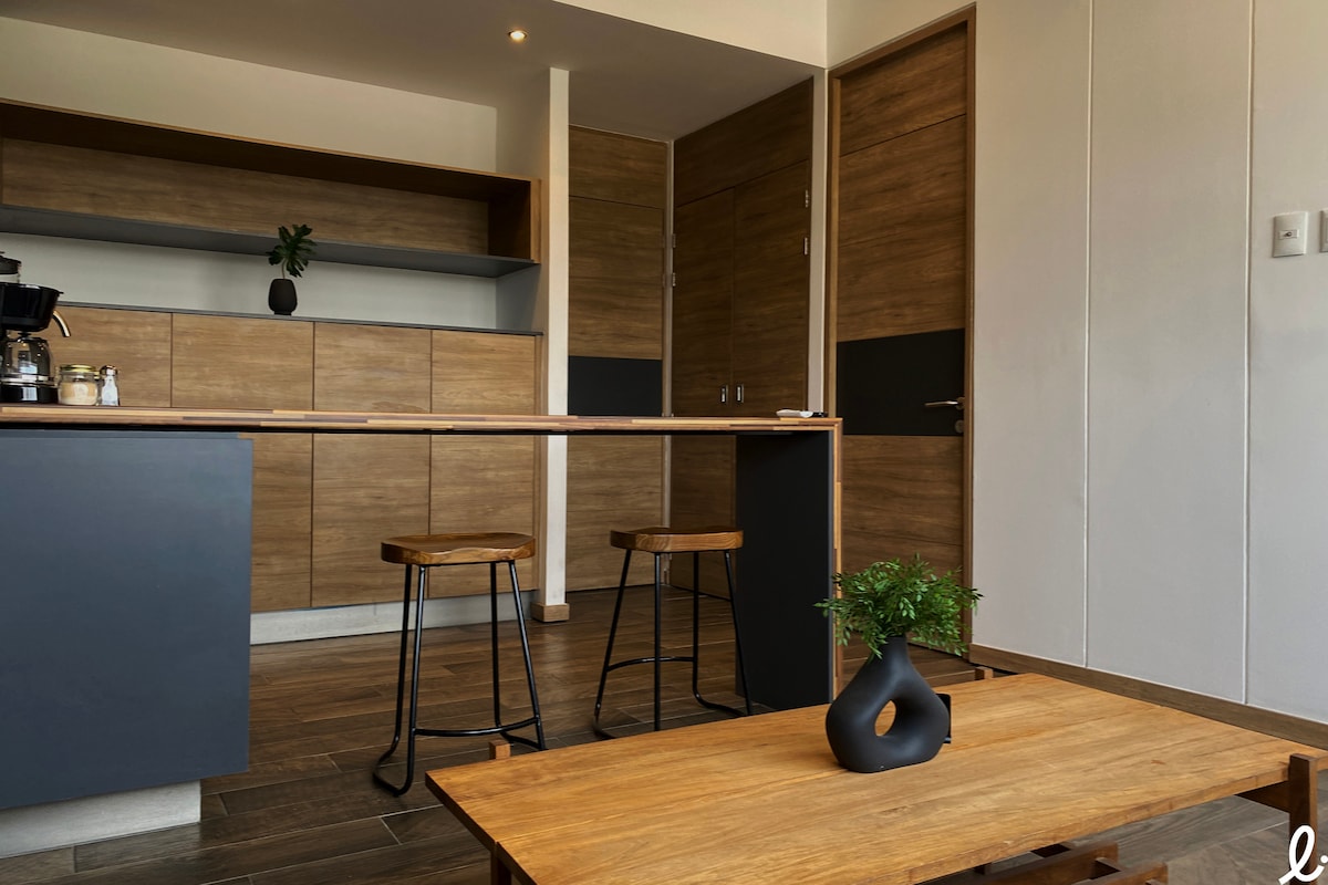 A modern kitchen area features wooden cabinetry and a dark countertop. Stools made of wood are positioned at the bar, alongside a simple wooden table. A small indoor plant is placed on the table, adding a touch of greenery to the space.