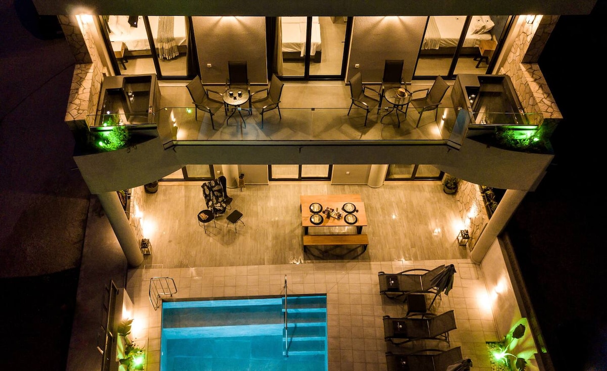 An aerial view captures the dual-level outdoor space featuring a clear swimming pool lit with ambient lighting. Multiple lounge chairs and small tables are arranged around the pool area, while large glass doors provide access to the interior of the building.