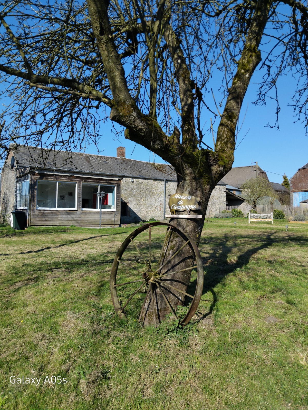 A weathered wooden wheel leans against a tree in a grassy yard. The house is visible in the background, featuring a simple design with a gray roof and large windows. Clear blue skies provide a bright backdrop, enhancing the rural setting.