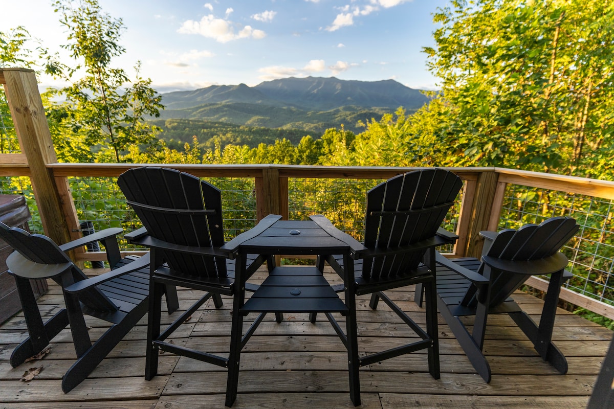 A wooden deck features four black Adirondack chairs arranged around a small side table, overlooking expansive mountain views. Lush greenery surrounds the area, with the sun illuminating the landscape and highlighting the serenity of the setting.