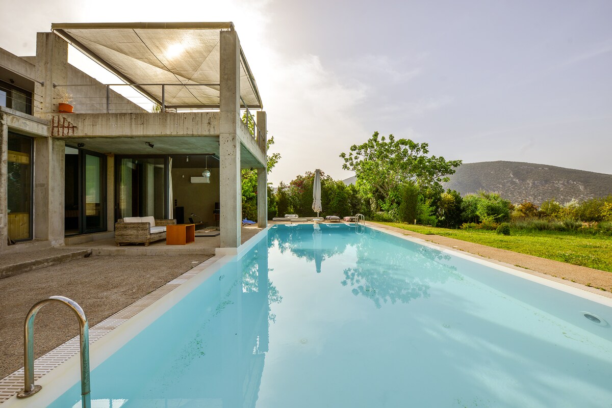 An outdoor area is showcased, featuring a private pool with clear water and a sunbathing space furnished with day beds. The villa's modern architecture is visible, with a shaded terrace that offers views of the surrounding greenery and hills in the background.