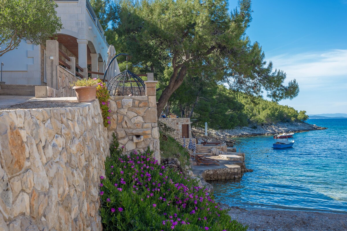 A stone pathway leads to the water's edge, bordered by blooming purple flowers. A small boat is visible on the calm bay, with greenery lining the shore, and clear blue skies above. The scenic coastal view enhances the natural beauty of the setting.