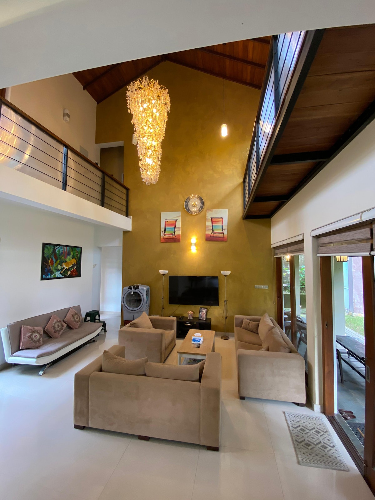 A spacious living area is showcased with a high ceiling and wooden beams. A modern chandelier hangs above, while comfortable sofas are arranged around a glass coffee table. An entertainment unit is visible along with a piece of wall art, enhancing the inviting space.