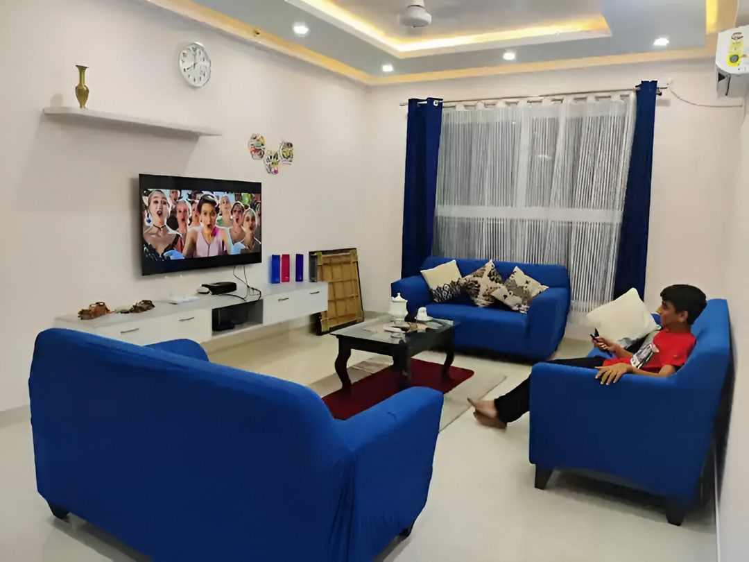 A cozy living area features two blue sofas arranged around a glass coffee table, while a flat-screen TV displays lively content. Natural light filters through sheer curtains, and a wall clock accents the space. A welcoming atmosphere is evident, ideal for family gatherings.