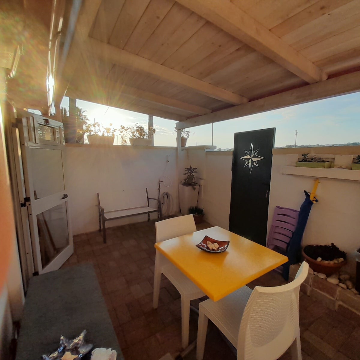 An outdoor space is presented with a yellow table and four white chairs. Green plants line the walls, and a door with a nautical star design is visible. Soft sunlight filters through the overhead structure, creating a warm ambiance in the area.