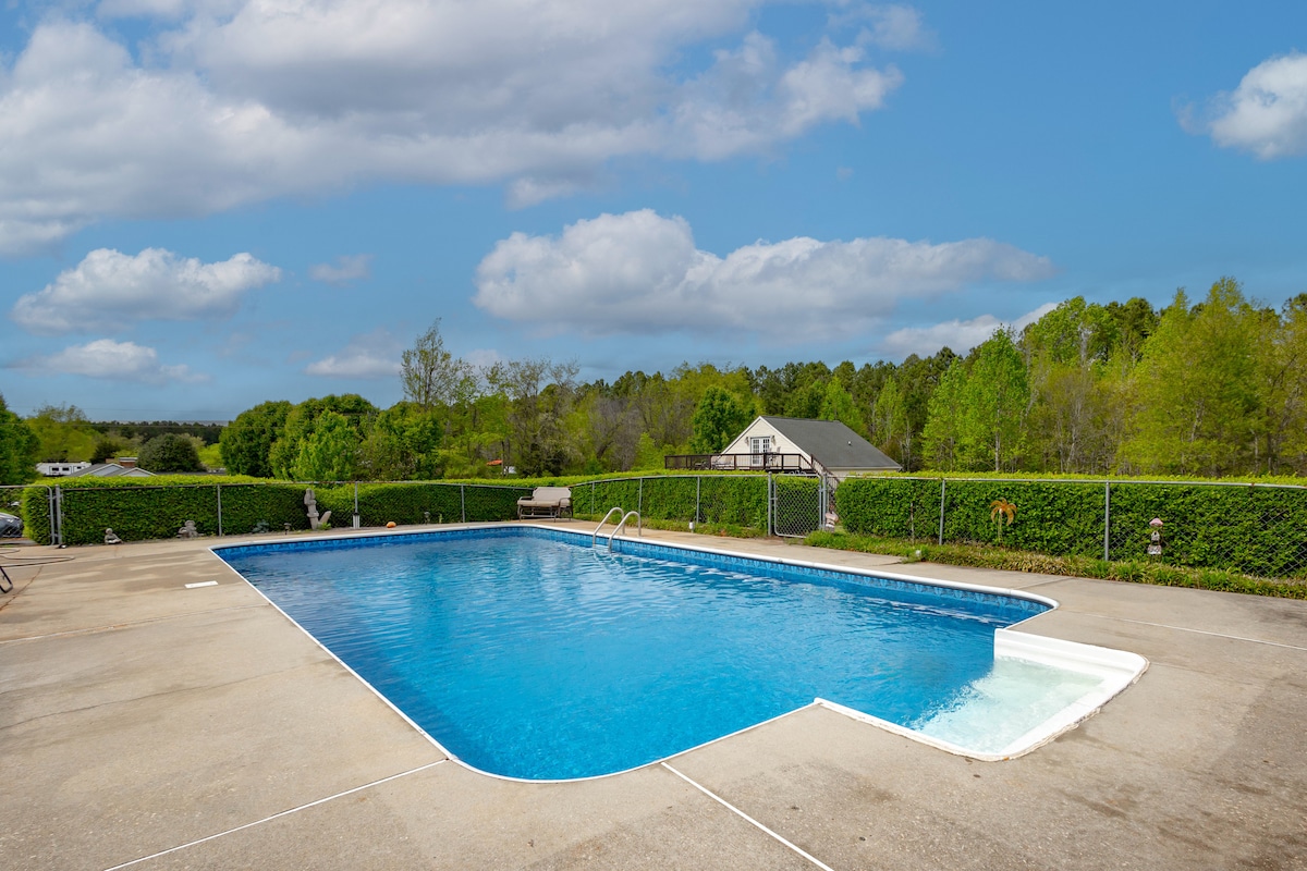 A seasonal pool is situated within a paved area, featuring clear blue water. Lush green hedges surround the pool, creating a private atmosphere. A shallow entry steps down into the pool on one side, inviting leisurely dips on warm days.