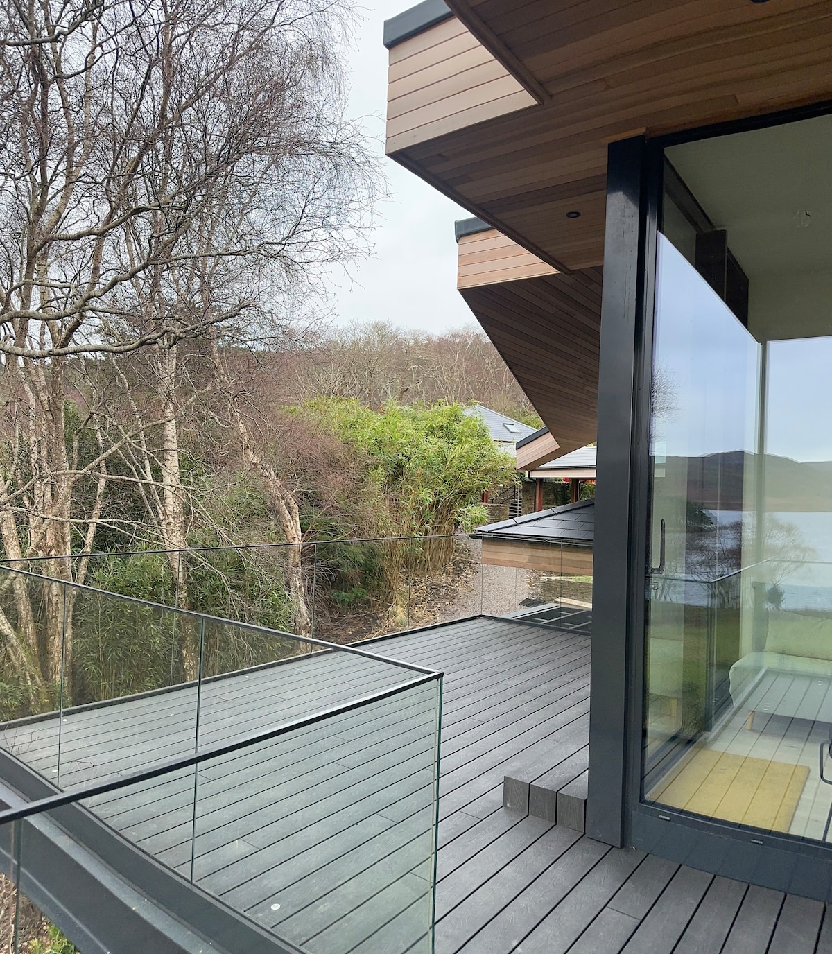 A wraparound balcony is featured, with a wooden deck and glass railing offering unobstructed views of the surrounding trees and landscape. The architectural design includes modern lines and a natural aesthetic, seamlessly connecting indoor and outdoor spaces.