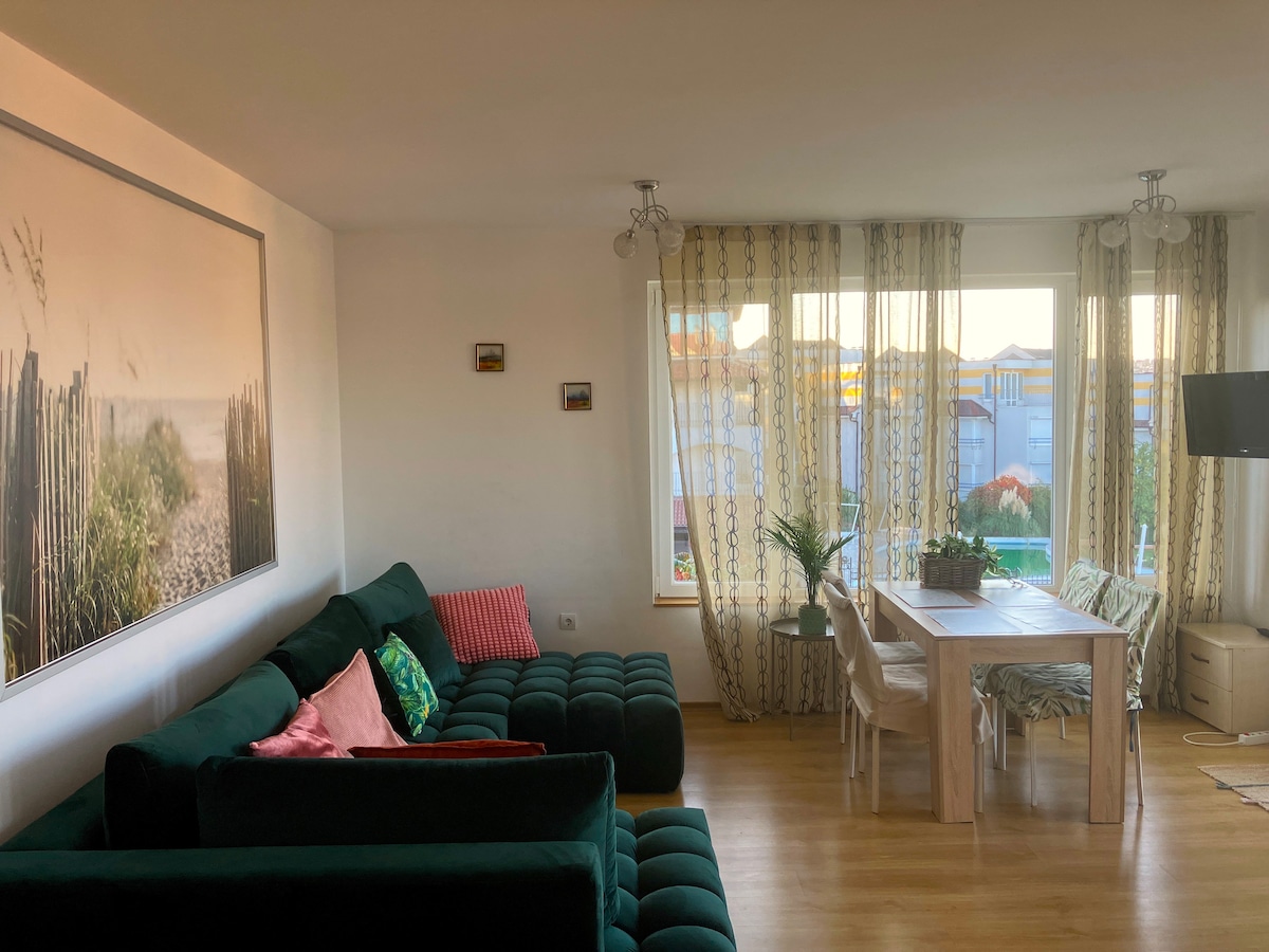 A spacious living room features a green sectional sofa with colorful pillows. A wooden dining table surrounded by chairs is positioned near large windows with sheer curtains, allowing natural light to brighten the area. A decorative plant adds a touch of greenery.