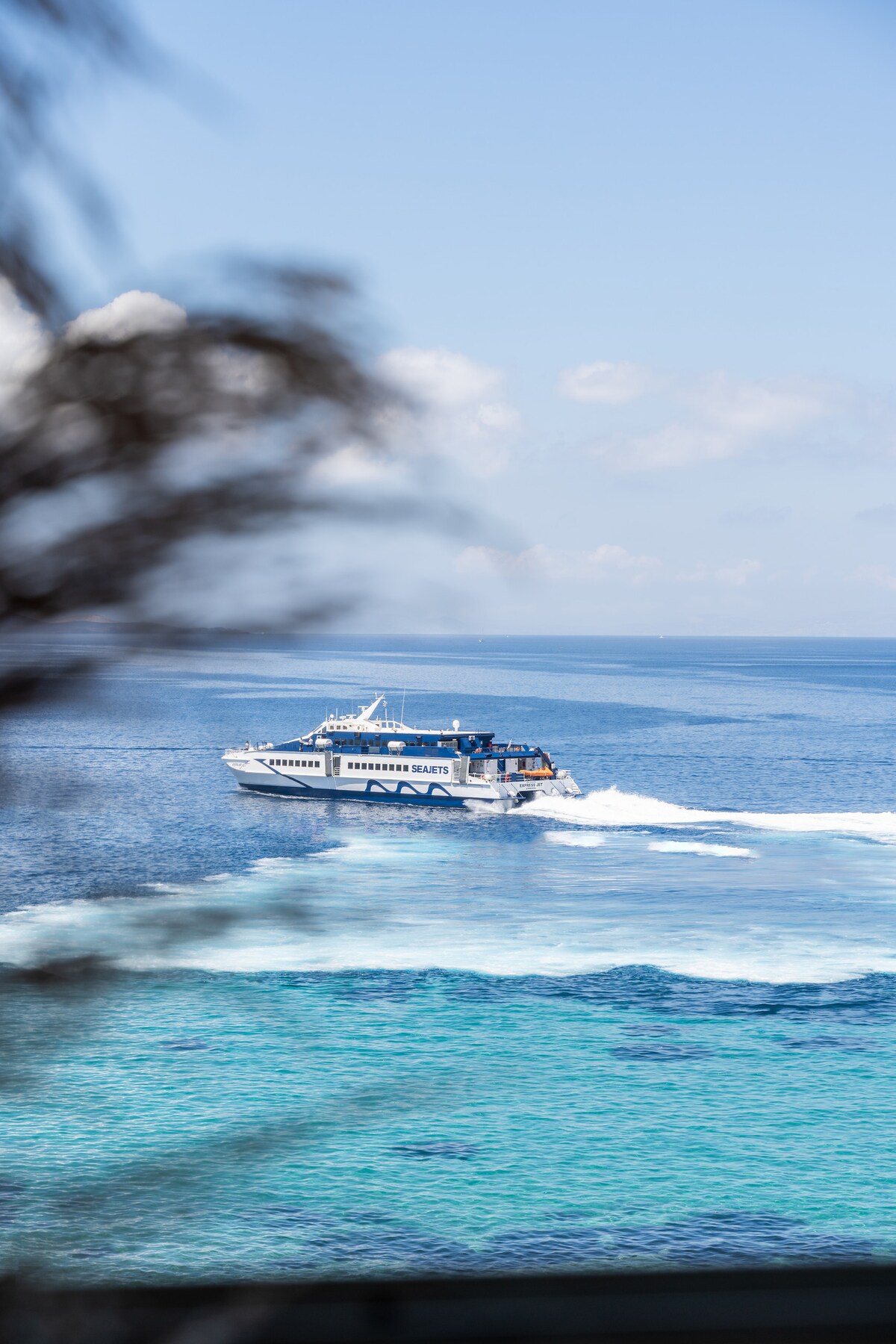 A ferry is seen gliding across the azure waters, leaving a frothy white trail behind. The scene captures the vibrant hues of the sea, transitioning from deep blue to lighter shades, under a clear sky with scattered clouds.