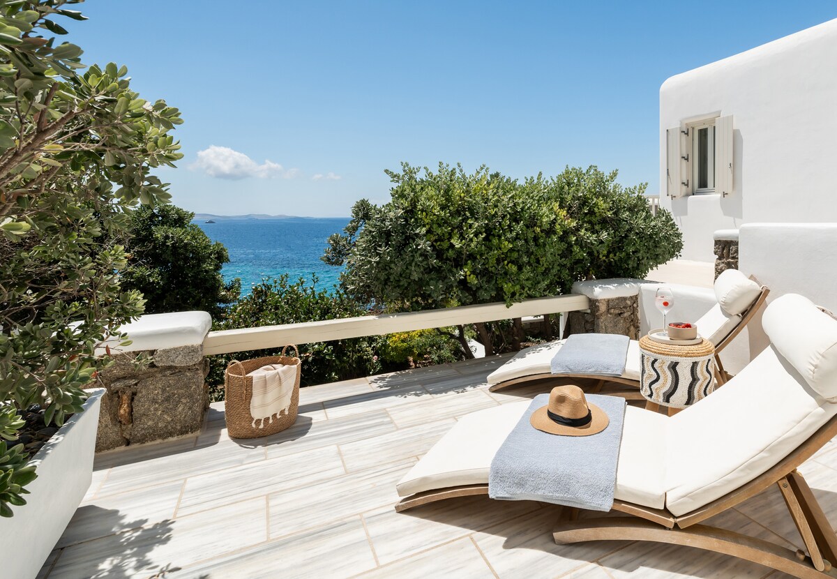 A private terrace is displayed with two sun loungers positioned for optimal sea views. Green foliage provides natural privacy, with a woven basket and decorative side table enhancing the space. The expansive blue sky complements the tranquil sea, creating a serene outdoor area.