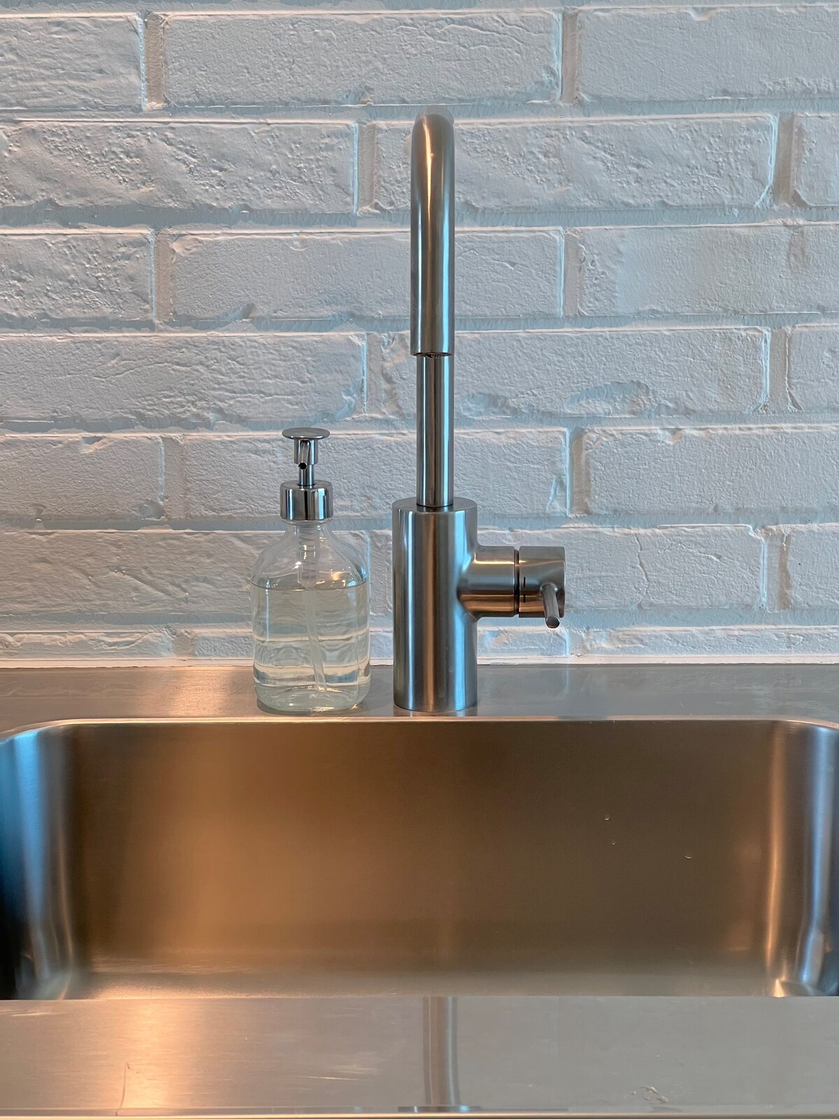 A stainless steel sink is positioned against a textured white brick wall. A modern faucet, featuring a sleek design, is positioned above the sink. A clear soap dispenser is placed next to the faucet, enhancing the minimalist aesthetic of the kitchen area.