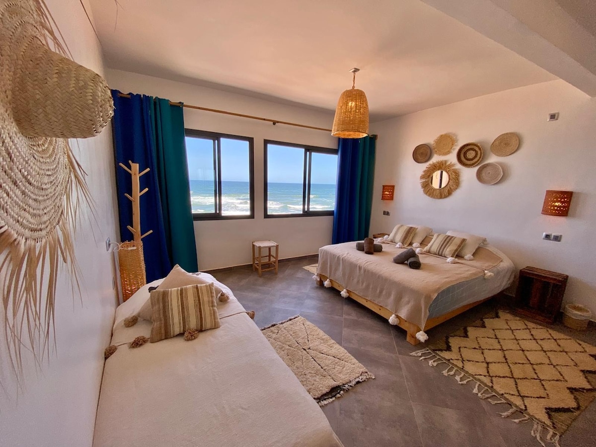 A spacious room features two large beds adorned with neutral bedding and decorative pillows. Natural light floods in through large windows, which provide views of the ocean. Woven wall decor and warm lighting elements enhance the inviting atmosphere, while a cozy seating area is present.