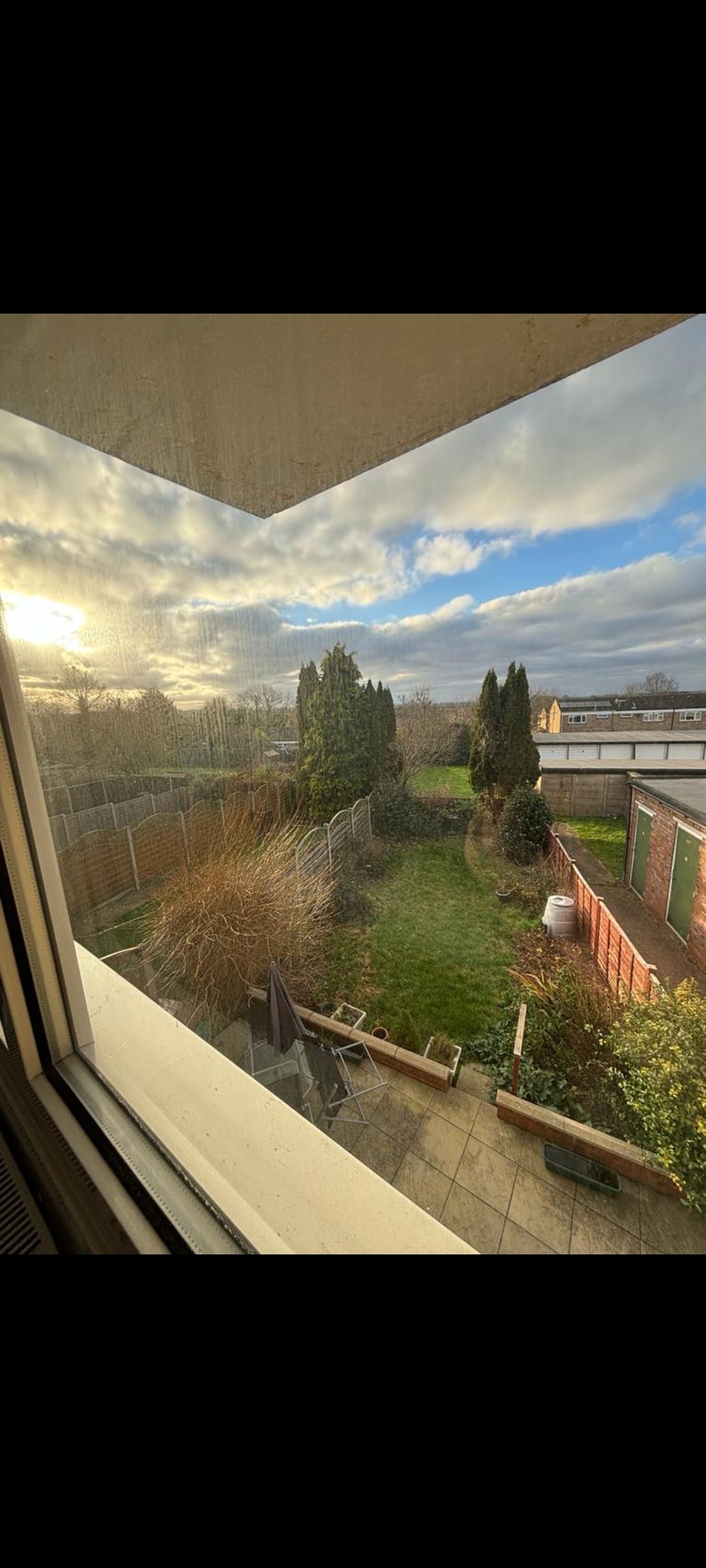 A view from a window captures a serene outdoor space featuring a well-maintained garden with green grass and shrubs. A patio area is visible, along with a few outdoor furniture pieces and a glimpse of neighboring structures against a cloudy sky.
