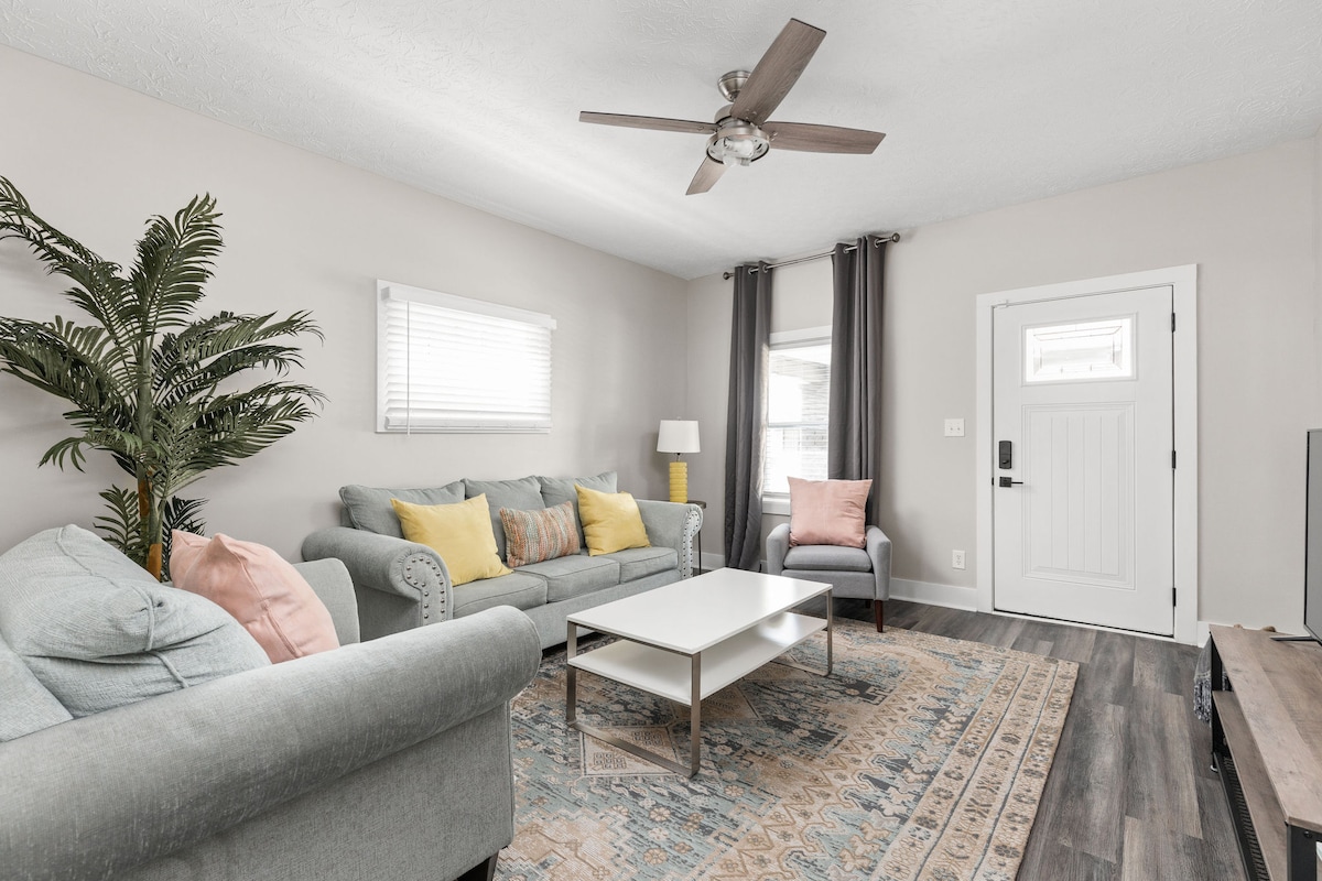 The living area features a light grey sofa and a matching loveseat accented with pastel-colored pillows. A coffee table sits in the center, complemented by a patterned area rug. Natural light enters through a window dressed with sheer curtains, and a ceiling fan provides airflow.