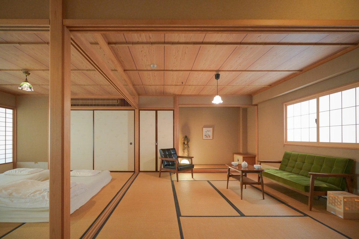 A spacious room features traditional tatami flooring, complemented by wooden beams on the ceiling. Two futons are neatly arranged on one side. A green sofa and a wooden chair are positioned around a low table, creating a cozy seating area. Natural light filters through sliding windows.
