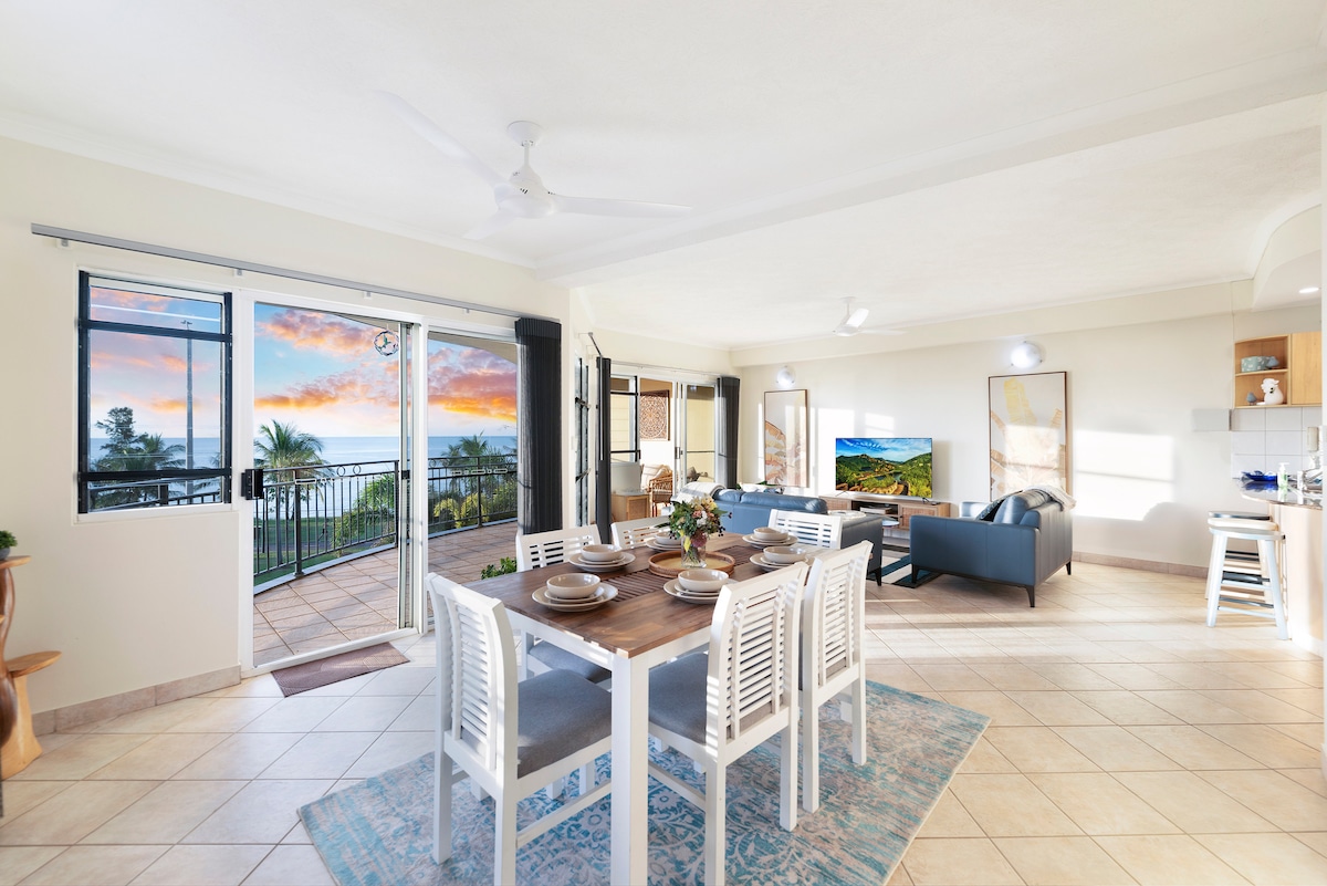 An open-plan living and dining area is observed, featuring a wooden dining table set for six, complemented by white chairs. Large windows offer views of a sunset over the sea. A comfortable sofa and a wall-mounted flat-screen TV are also present, enhancing the inviting space.
