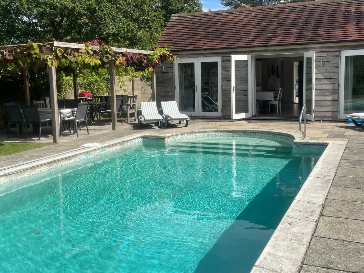 A private heated swimming pool is surrounded by a paved patio and a lush garden. Relaxation areas feature sun loungers and an outdoor dining space under a pergola, which is adorned with greenery. The cottage's large glass doors open directly onto the pool area.