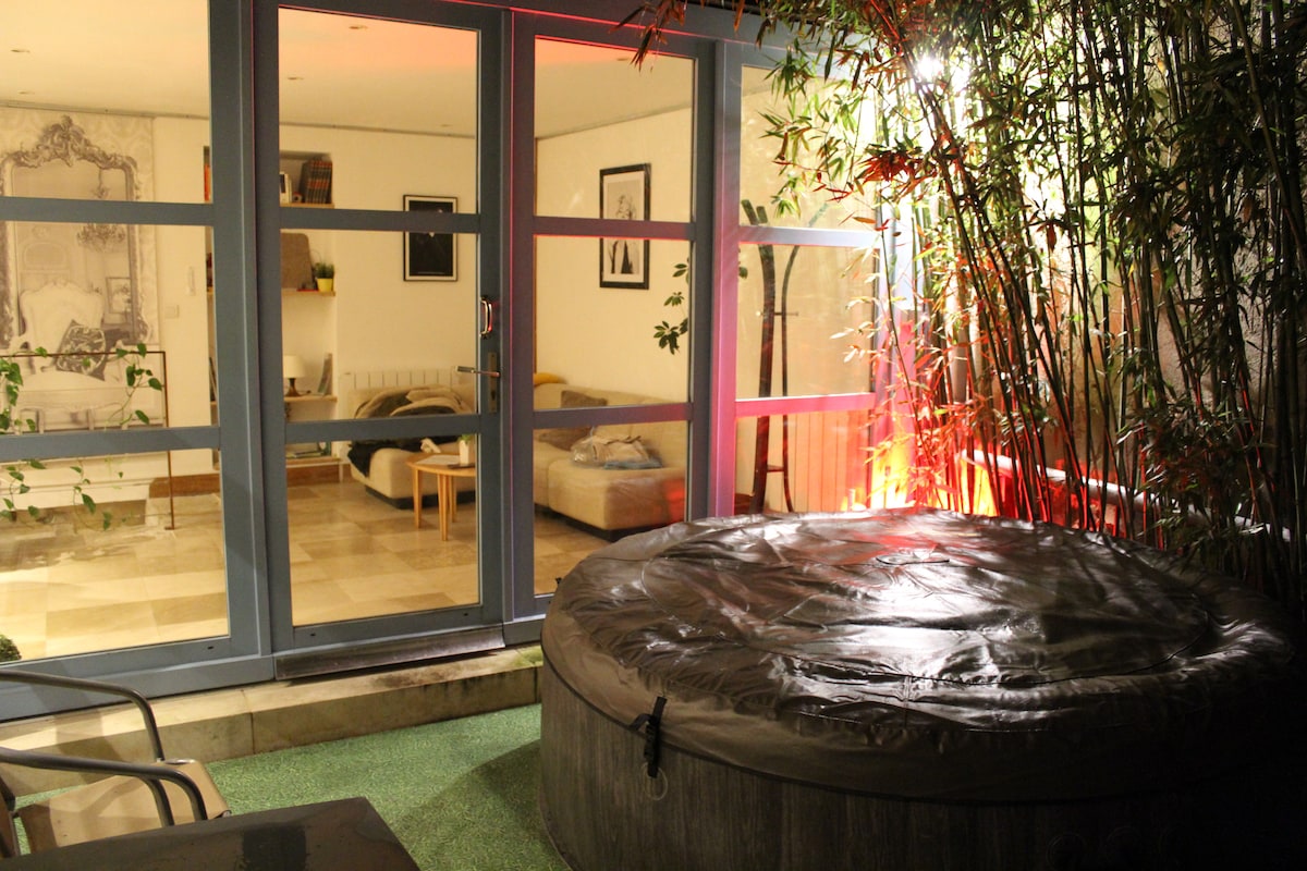 A secluded patio area features a round hot tub, surrounded by bamboo plants. Large glass doors lead to a softly lit interior space, showcasing a comfortable seating area with neutral-toned furnishings. The overall atmosphere is quiet and intimate, ideal for relaxation.