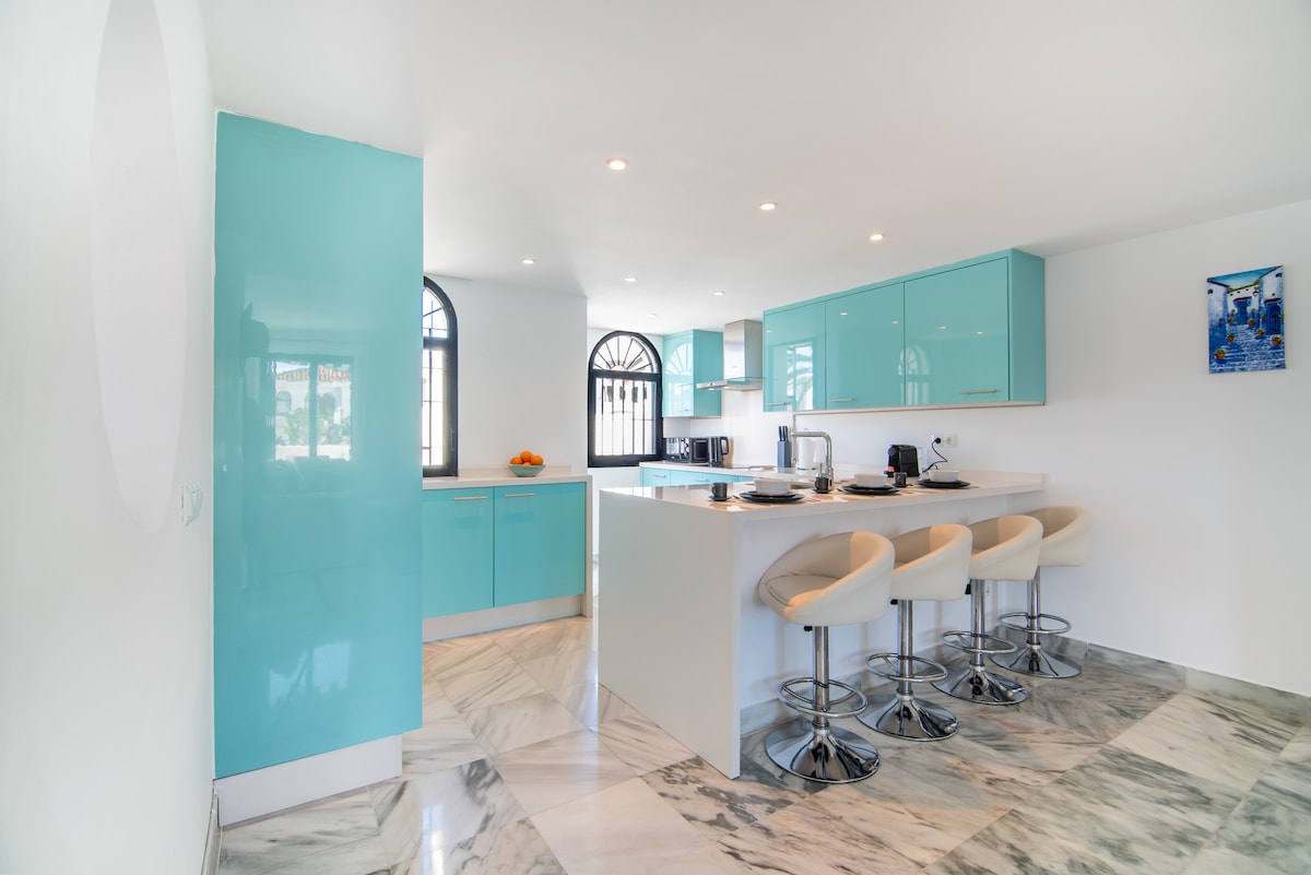 A modern kitchen features turquoise cabinetry and marble flooring. Bar stools are positioned around a white countertop, with natural light entering through large windows. A small dining area is visible, adding functionality to the open-space design, creating a bright and inviting cooking environment.