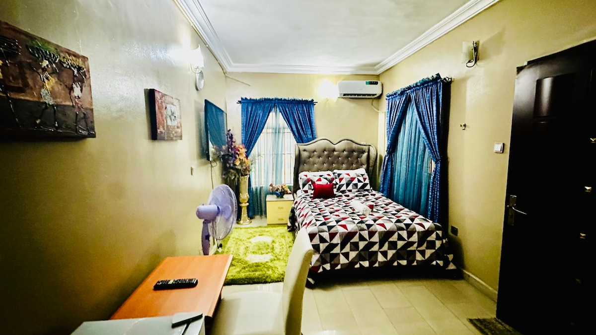 A cozy bedroom features a patterned bedspread on a spacious bed, set against a soft beige wall. Blue curtains frame the window, allowing natural light to fill the room. A small desk and chair provide a work area, while a fan and television add to the amenities.