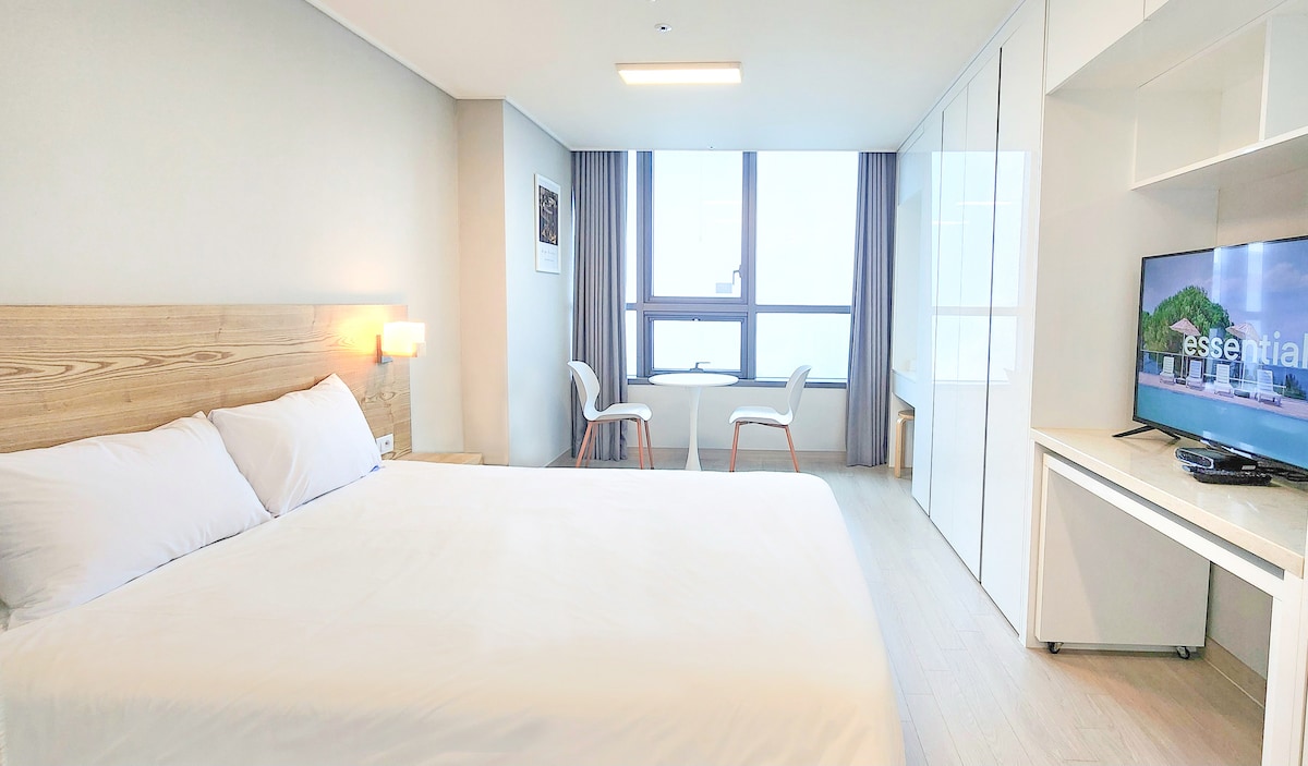 A well-lit bedroom features a spacious bed with crisp white linens, flanked by a bedside lamp. Two chairs are positioned near a window that offers natural light, with a contemporary TV mounted on a white console against the wall.