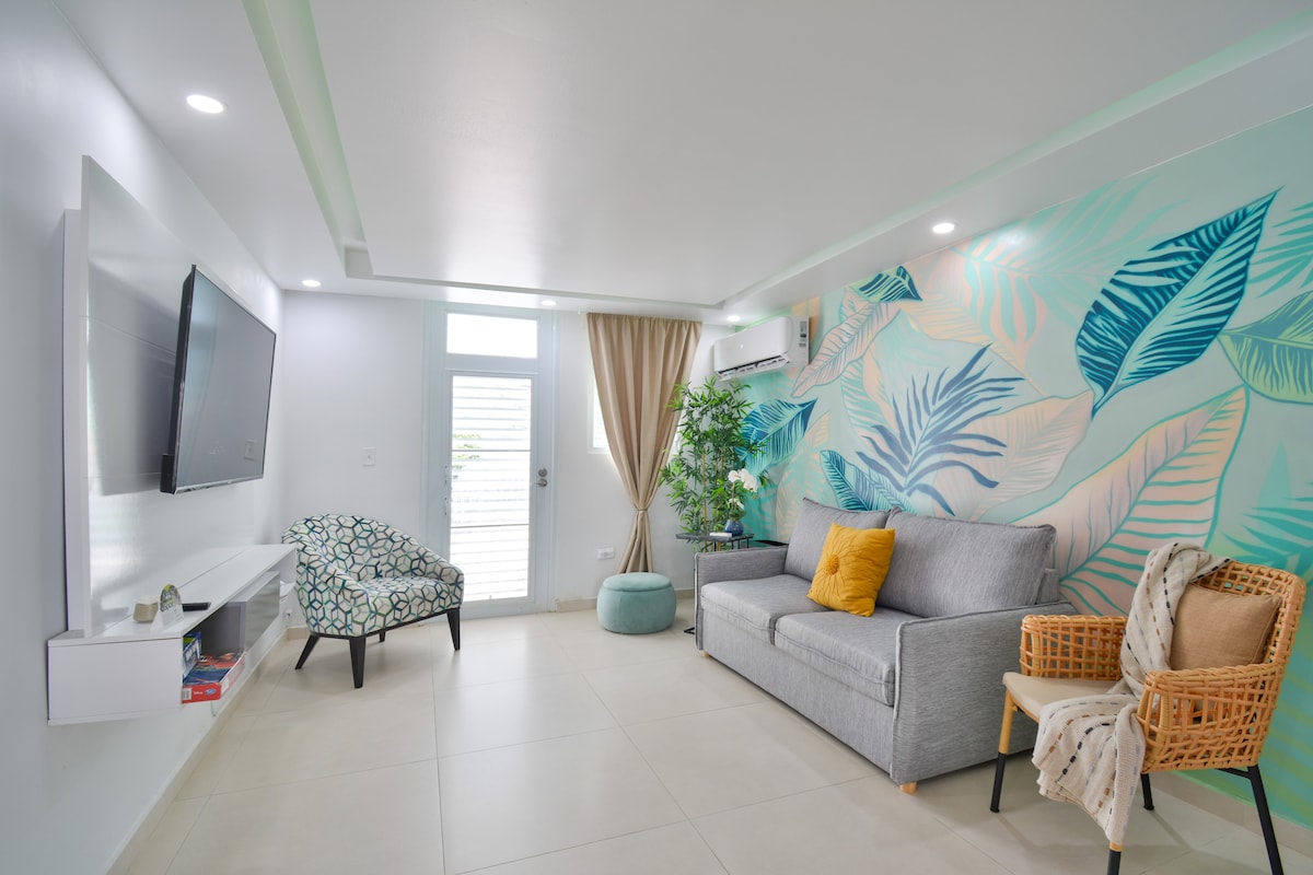 A modern living area is characterized by light tile flooring and a fresh green accent wall featuring leaf patterns. A neutral-colored sofa is complemented by a vibrant yellow cushion. Soft natural light enters through a door leading to the outside, and two chairs provide additional seating.