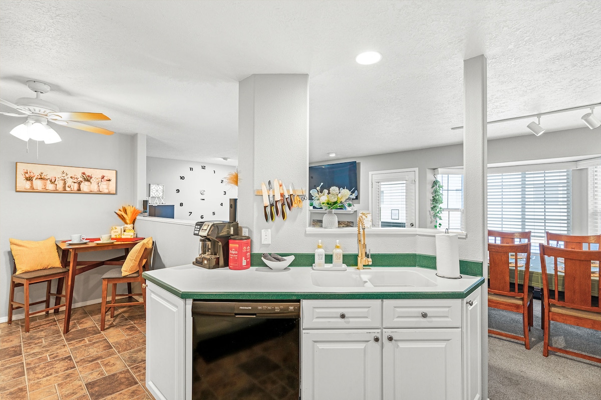 The spacious kitchen features modern appliances and a large countertop with a sink. A dining area is visible in the background, showcasing a wooden table with yellow-accented chairs. Natural light filters through the nearby window, enhancing the bright and open space.
