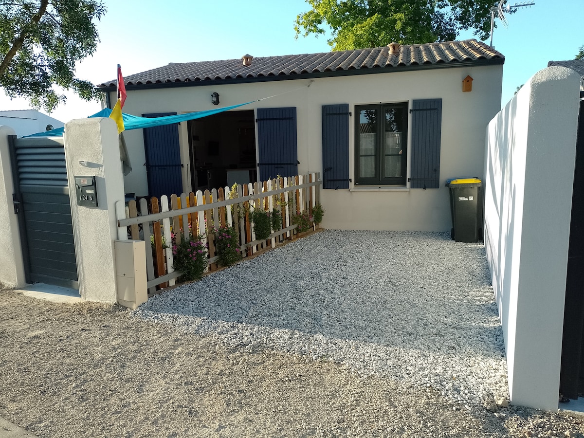 A charming exterior of a single-story house is depicted, featuring light-colored walls and blue shutters. A gravel surface is present, bordered by a wooden fence adorned with flowers and greenery. A mailbox and a green waste bin are visible, enhancing the inviting curb appeal.