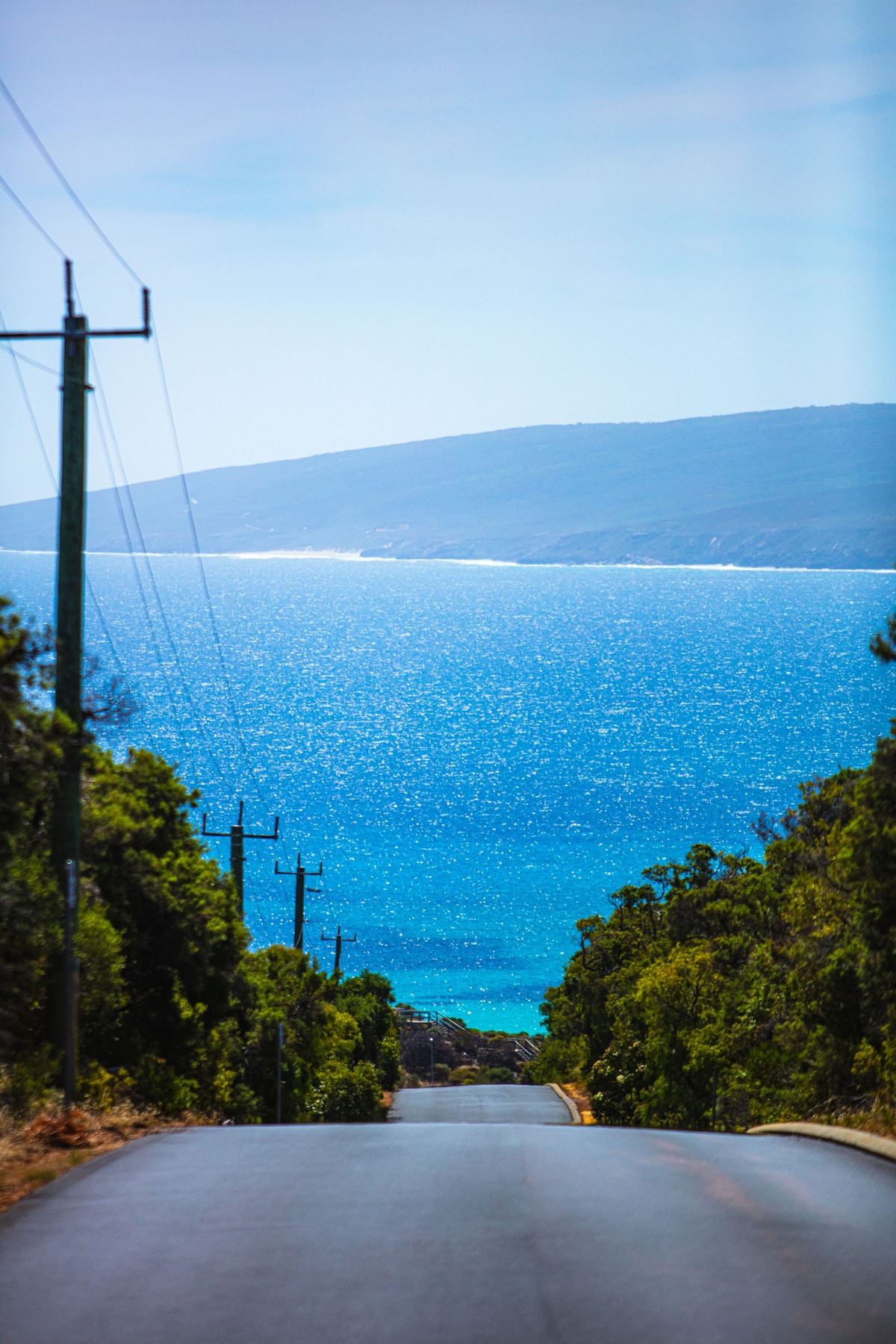 A road leads toward the ocean, framed by lush greenery on either side. The water displays a vibrant blue hue, reflecting sunlight, while distant hills are visible along the coastline. Telephone poles line the road, subtly guiding the viewer's gaze toward the sea.