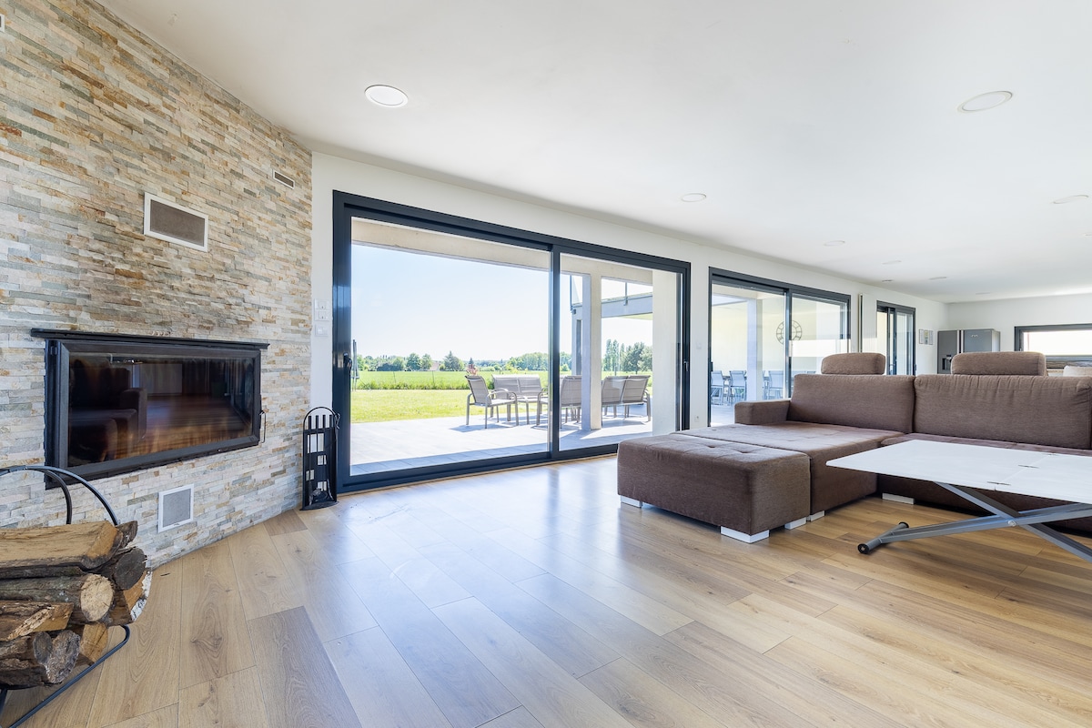 A spacious living area is showcased with large glass doors that provide views of the green landscape outside. A modern sofa is positioned near a stone fireplace, contributing to the inviting ambiance. Natural light illuminates the warm wooden floors, enhancing the open design.