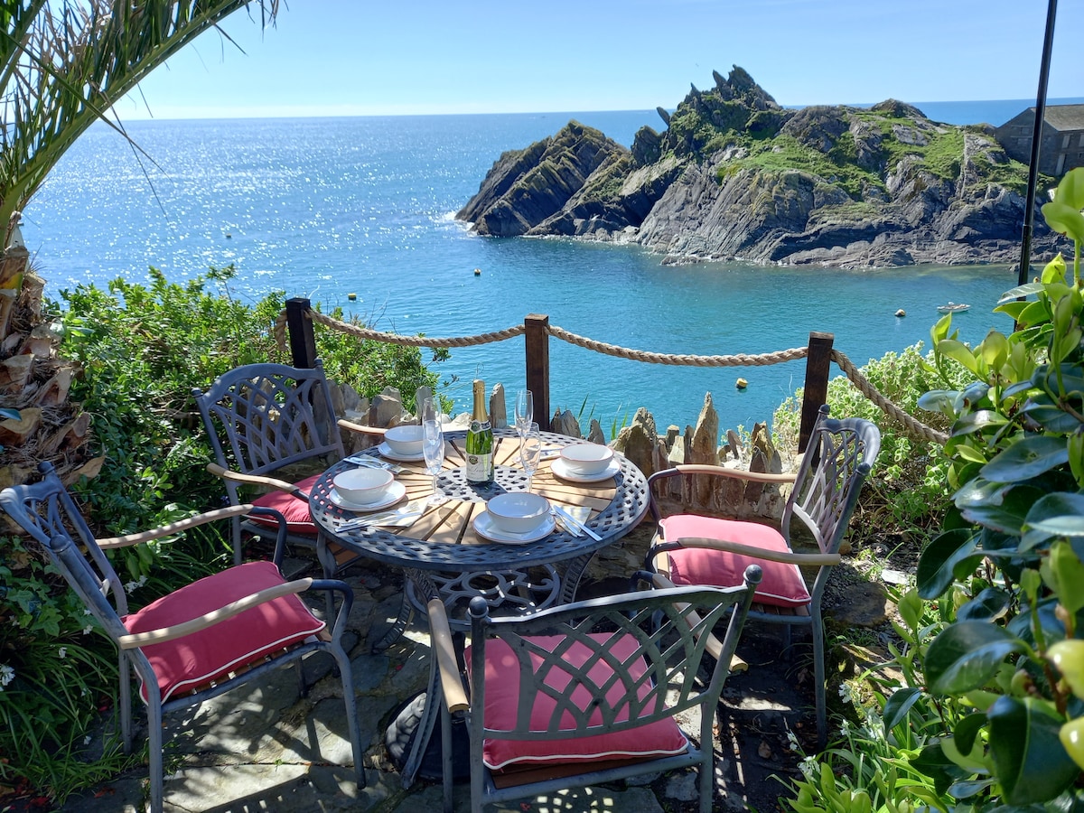 A circular garden table with four chairs is set for an outdoor meal, featuring plates and glasses. The stunning view of the coastal rock formations and sparkling sea complements the tranquil atmosphere, surrounded by lush greenery and palm-like plants.