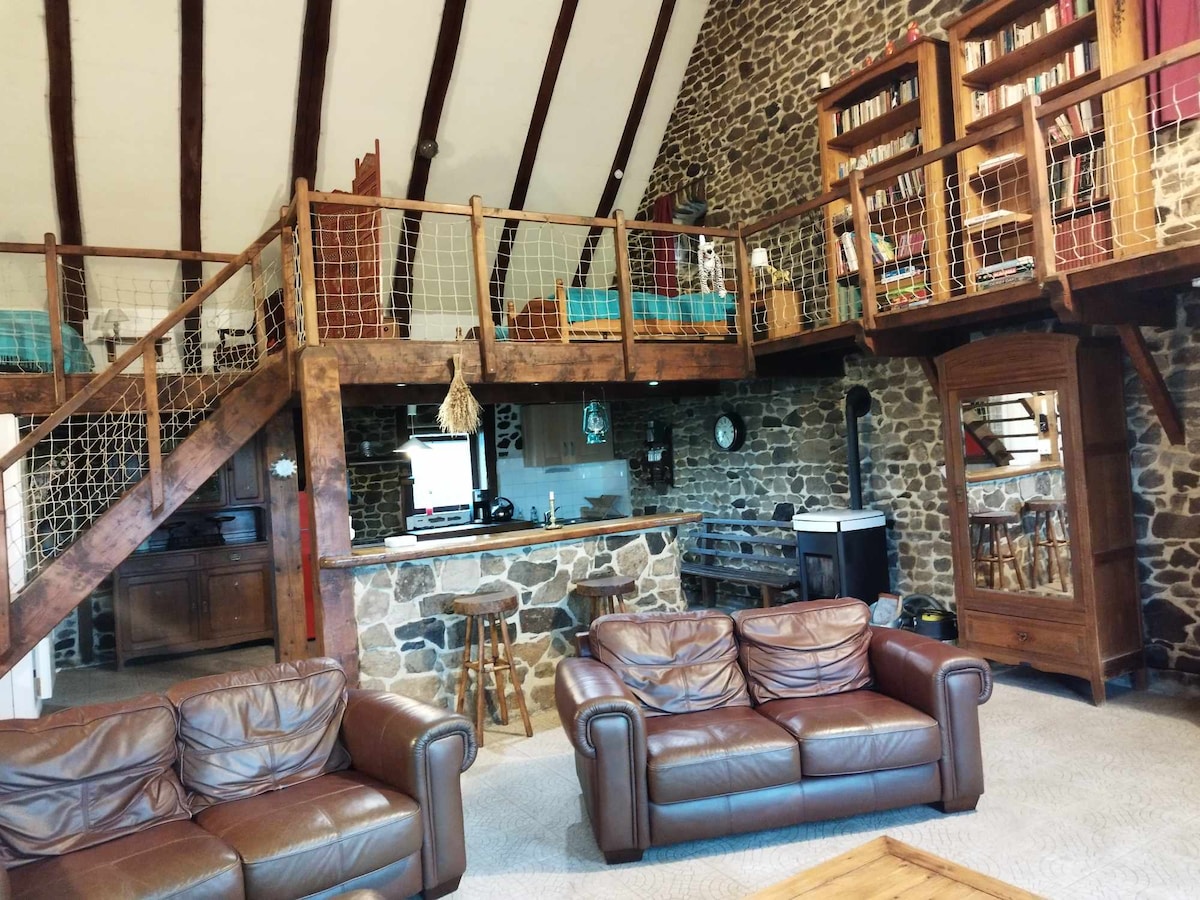 A spacious loft with rustic charm features exposed stone walls and high ceilings. A cozy seating area is arranged with leather sofas. A mezzanine overlooks the main level, showcasing additional sleeping space. The kitchen is visible, equipped with essential appliances, creating a harmonious living area.