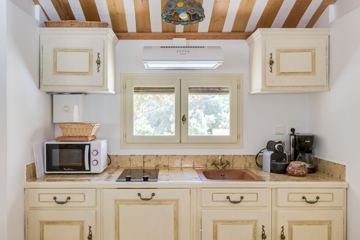 A well-equipped kitchen features cream-colored cabinetry and natural wood beams overhead. A microwave and coffee machine are visible, alongside a sink and stovetop integrated into the counter. Natural light enters through a window, offering a view of the outdoors.