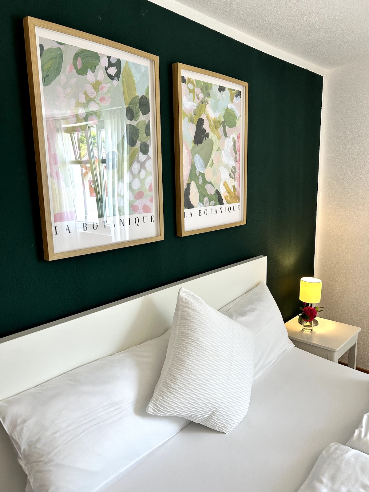 The bedroom features a spacious bed adorned with crisp white linens and a decorative pillow. Two framed botanical prints add color to the dark green wall, while a small lamp with a floral accent stands on a bedside table, providing soft illumination.