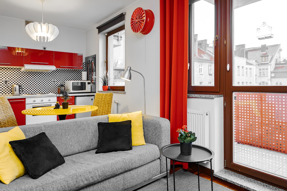 A modern living area features a grey sofa adorned with black and yellow cushions. A round black table stands nearby, and a well-lit kitchen with red cabinetry is visible in the background. Window coverings consist of bright red curtains, complementing the overall design.